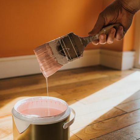 Mano sumergiendo un pincel en una lata grande de pintura rosa mientras gotea de nuevo en la olla.