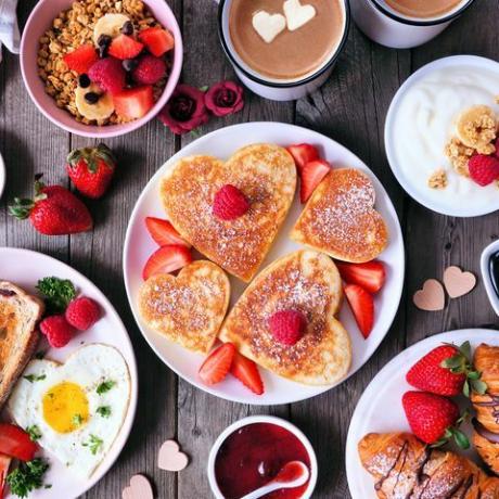escena de la mesa de desayuno del día de san valentín o de la madre sobre un fondo de madera oscura con panqueques en forma de corazón, huevos y comida temática de amor