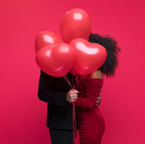 Una pareja joven vestida con ropa elegante besándose detrás de un montón de globos en forma de corazón.